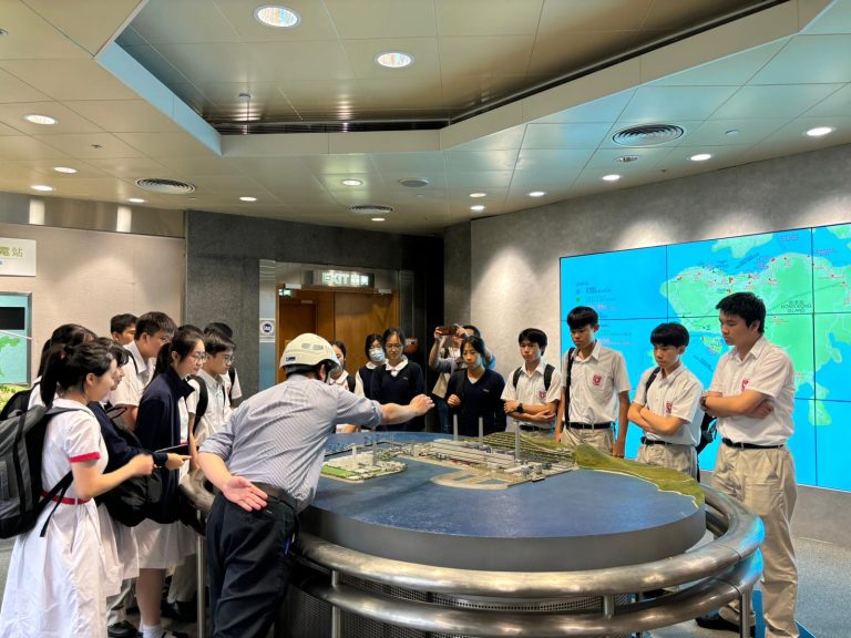 Students observing a large-scale model of a power station with an instructor gesturing towards it.