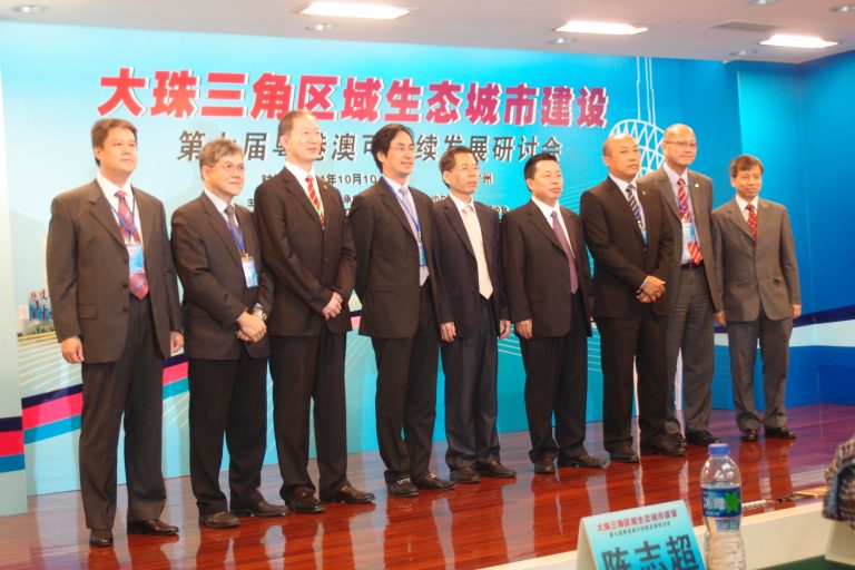 Seven men in suits standing on stage at a conference on eco-city development.