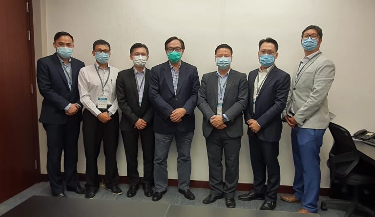 Seven men in business attire and face masks posing for a group photo indoors.