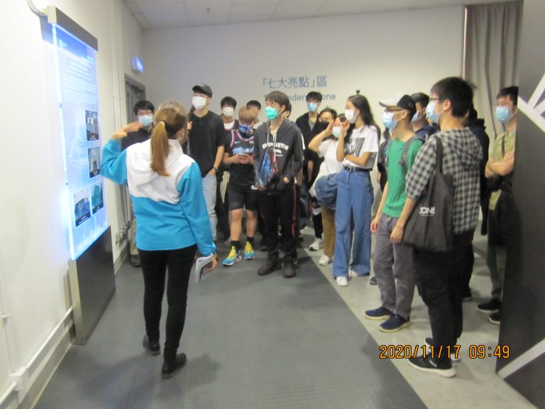 Group of people wearing masks listening to a presenter at an interactive display.