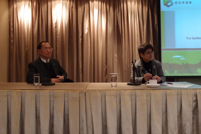 Two individuals seated at a table with microphones during a presentation.