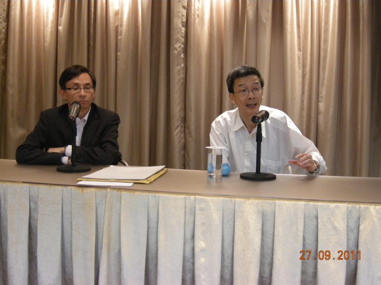 Two men seated at a table with microphones, one speaking, one listening, dated 27.09.2011.