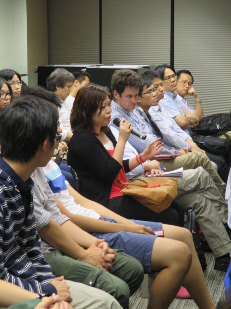 Audience members seated in a conference room, one woman speaking into a microphone.