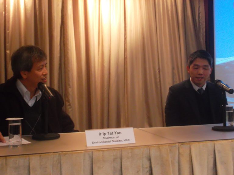 Two men speaking at a conference table with microphones and nameplate.