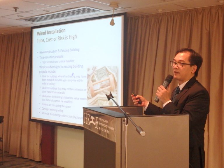 Man presenting at podium with microphone, behind screen showing wired installation slide.