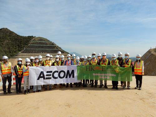 Group of construction workers in hard hats and safety vests holding AECOM banners at a worksite.