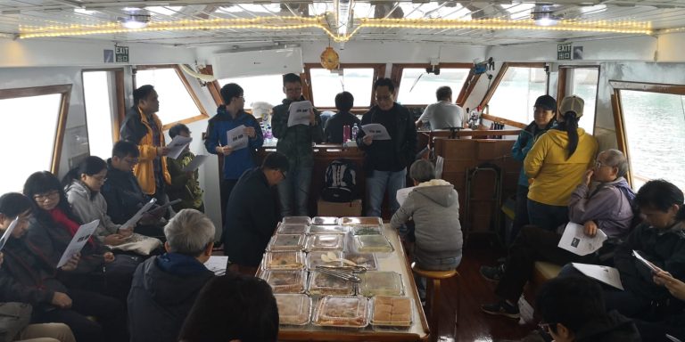 Group of people on a boat, some seated, some standing, reviewing papers near a table with food containers.