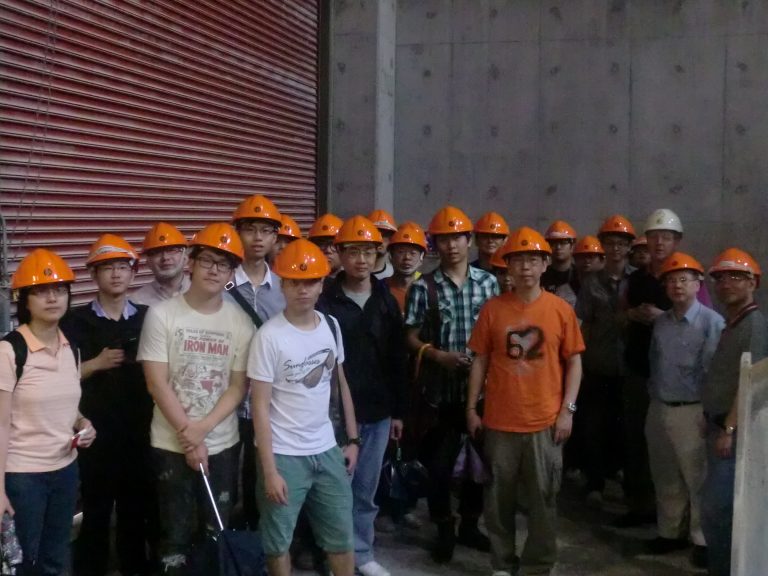 Group of people wearing orange hard hats posing for a photo at a construction site.