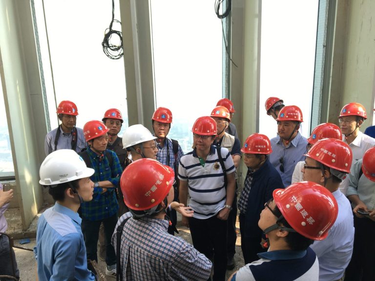 Construction workers in hard hats gathered on a high-rise building site, listening to a presentation.