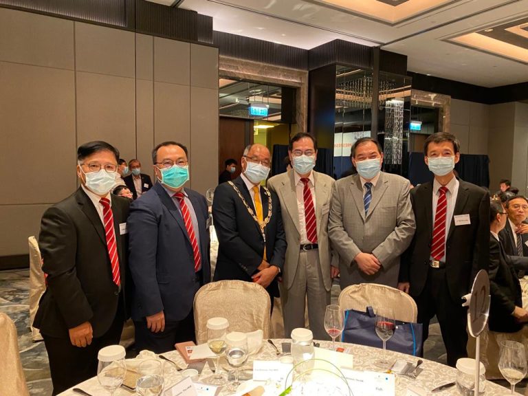 Six men in suits and face masks pose together at a formal event around a dining table.