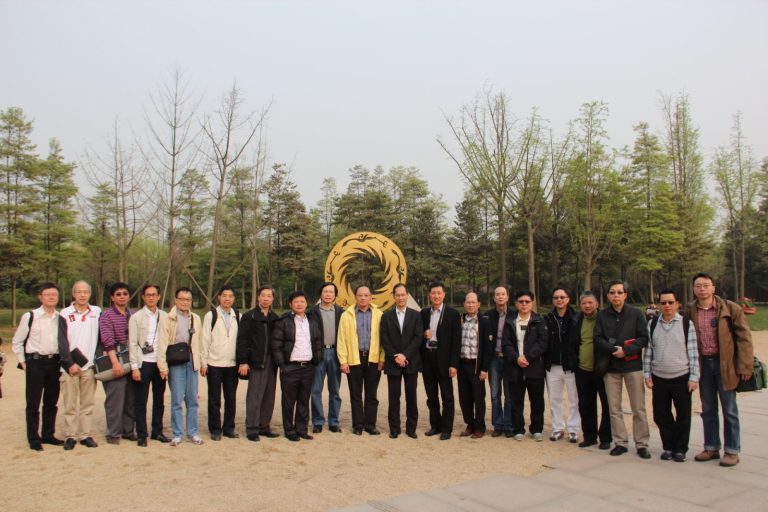 Group of men posing outdoors in front of a large golden circular sculpture with trees in background.