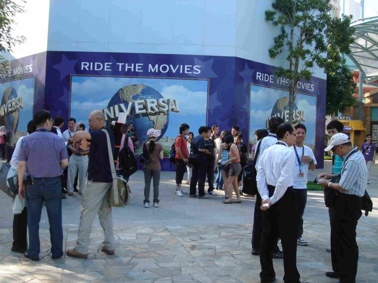 "People gather outside Universal Studios near a ""Ride the Movies"" attraction sign."