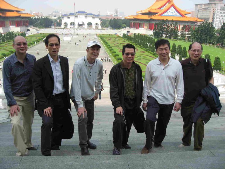 Six men posing together on steps in front of a large public square with traditional architecture.