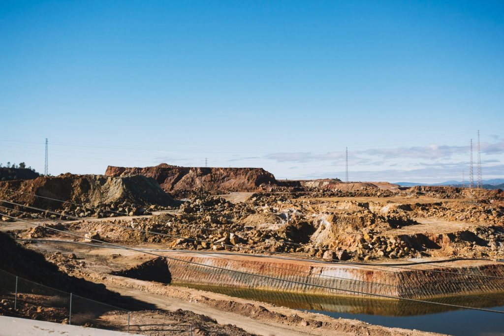 Open-pit mine landscape with excavated earth, power lines, and a small water body under clear blue sky.