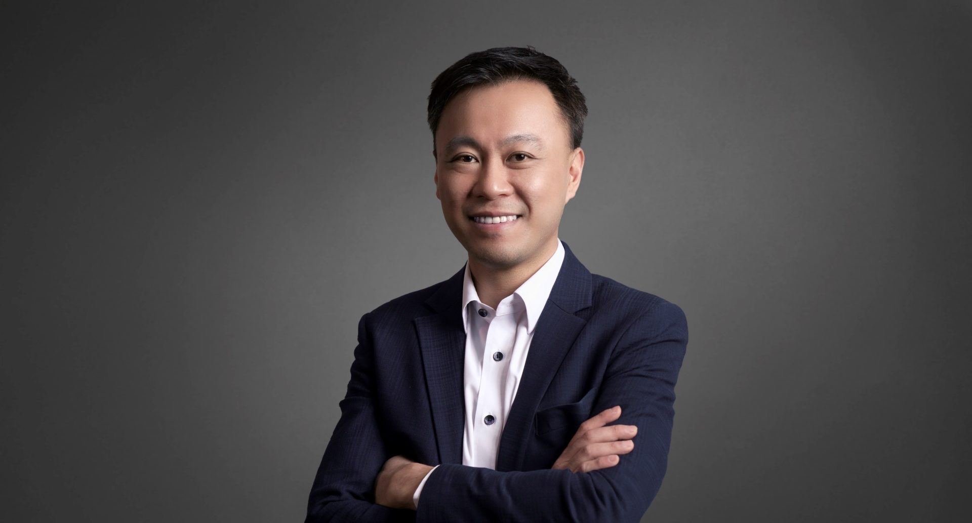 Professional portrait of a smiling East Asian man in a dark suit and white shirt with arms crossed.