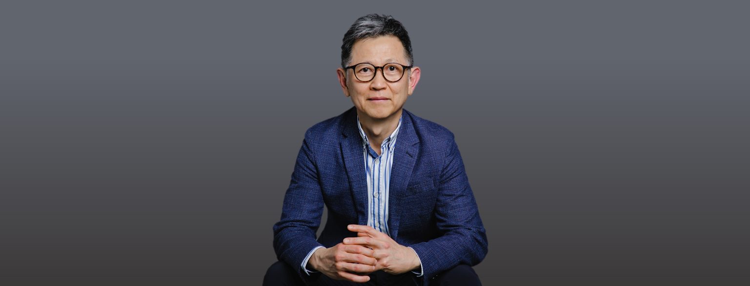 Man with glasses wearing blue blazer and striped shirt, seated with hands clasped.