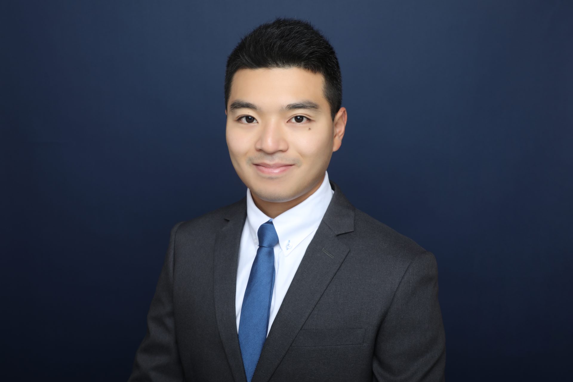Professional headshot of a man in a suit and blue tie against a dark blue background.