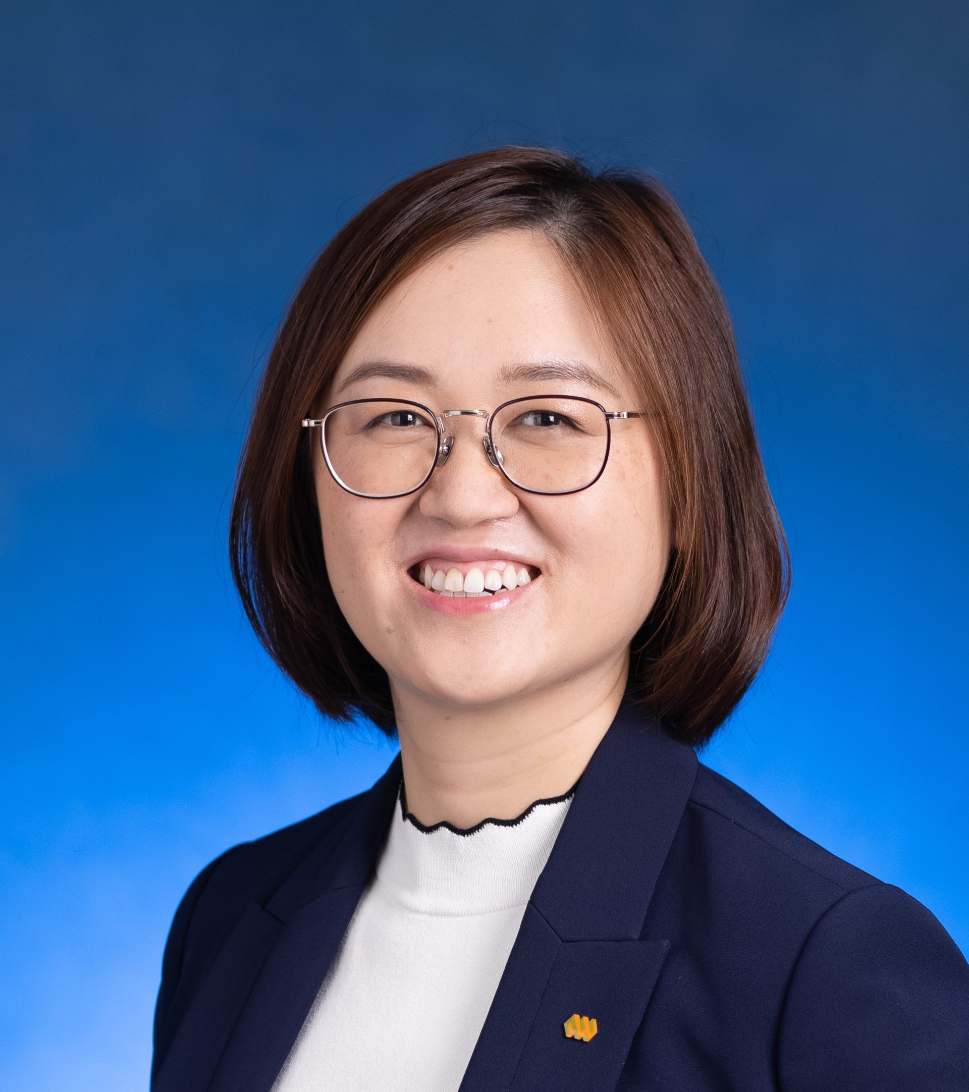 Smiling woman with glasses wearing a dark blazer over a white top against a blue background.
