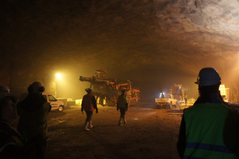 Construction workers in hard hats and vests inside a dimly lit tunnel with heavy machinery.