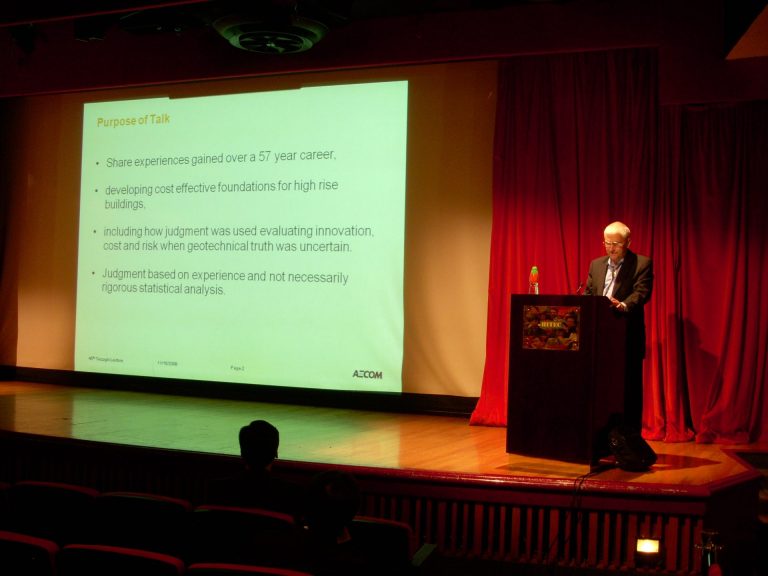 Speaker presenting at podium with slide on stage showing purpose of talk.