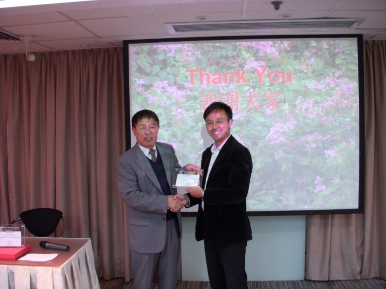 "Two men shaking hands while presenting an award on stage in front of a ""Thank You"" projection."