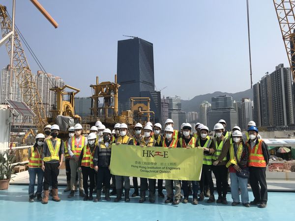 Construction workers in safety gear pose with HKIE banner at urban construction site.