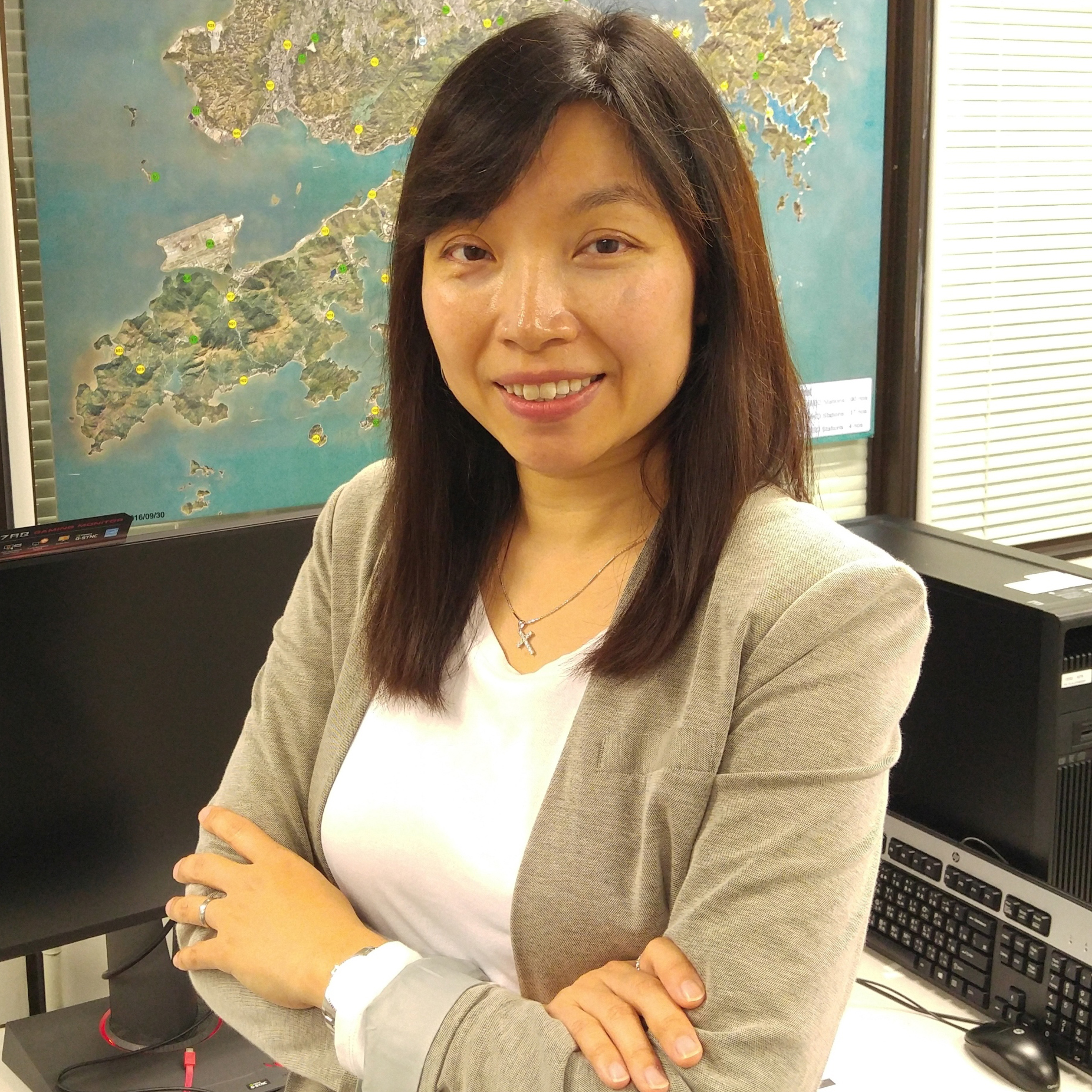 Smiling woman in office with arms crossed, map visible behind monitor