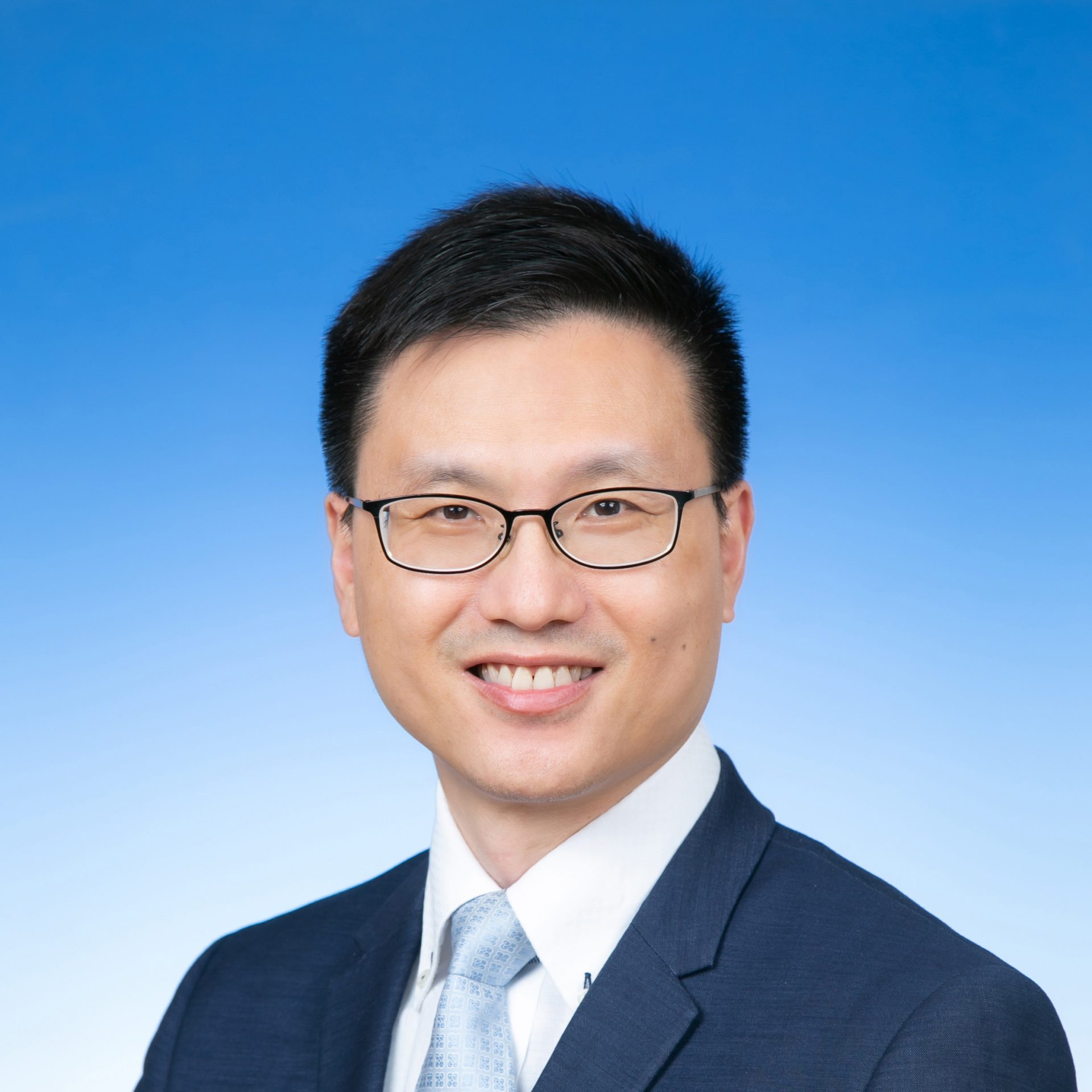Professional headshot of a smiling man wearing glasses, suit, and tie against a blue gradient background.