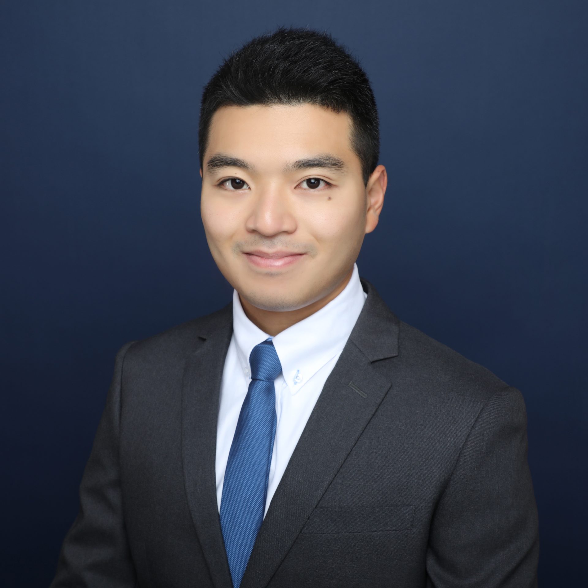 Professional headshot of a man in a gray suit and blue tie against a dark blue background.