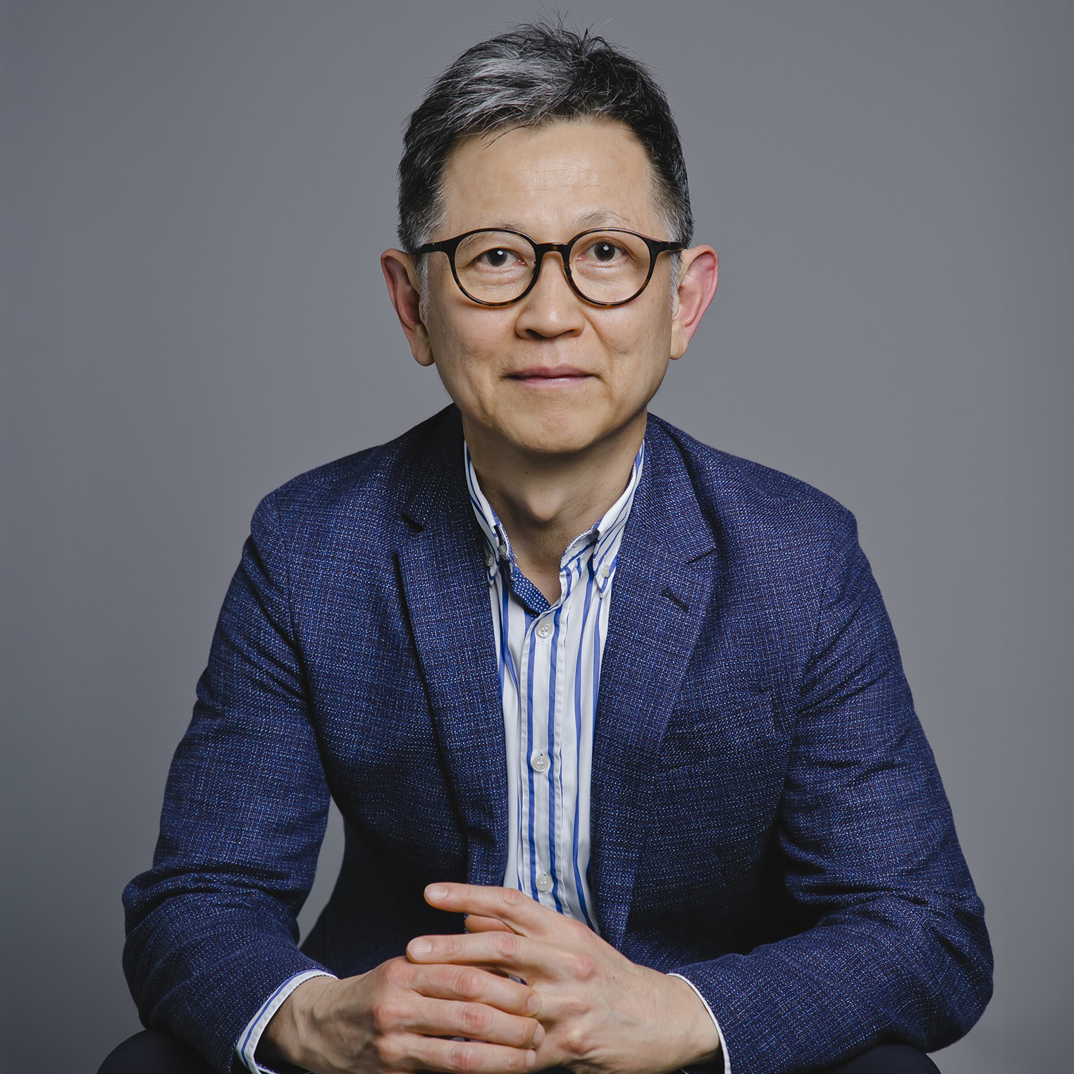 Man with glasses wearing blue blazer and striped shirt, hands clasped