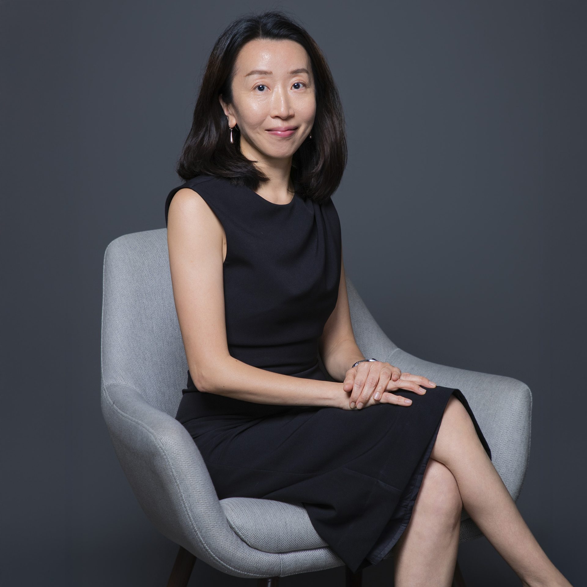 Professional portrait of a woman in a black dress seated in a gray chair.