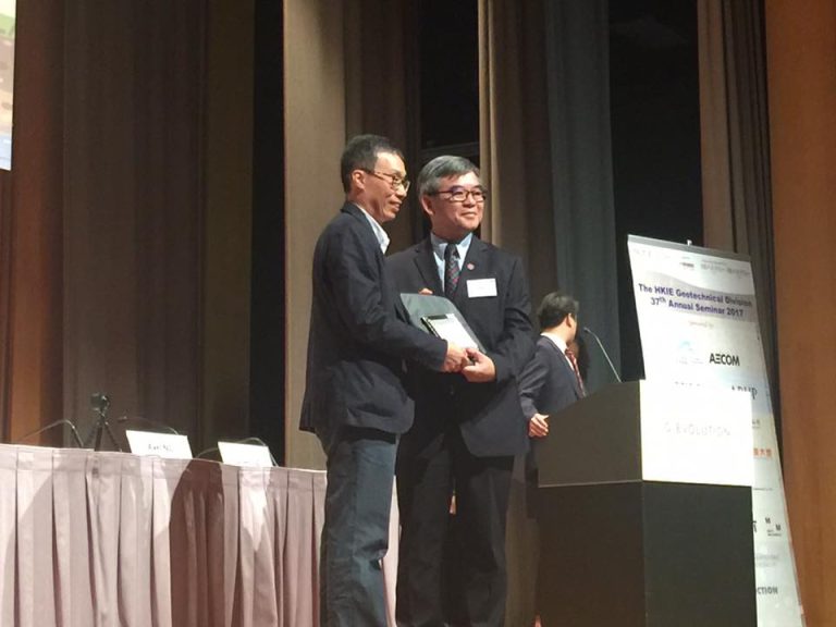 Two men in suits on stage, one presenting an award to the other at a seminar.