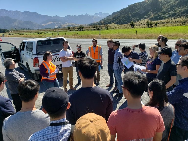 Group of people gathered outdoors near a white truck with mountains in background.