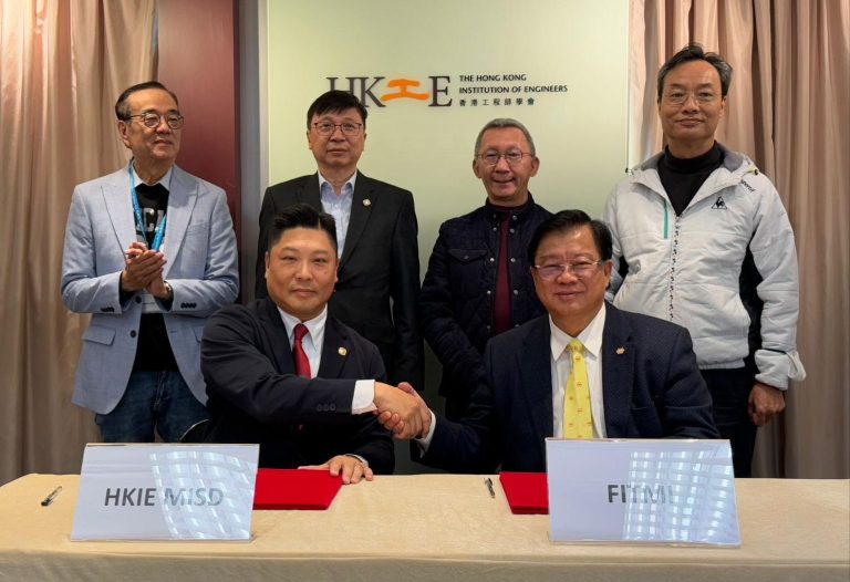 Two men shaking hands at a signing table with HKIE and FITMI nameplates, five others standing behind.