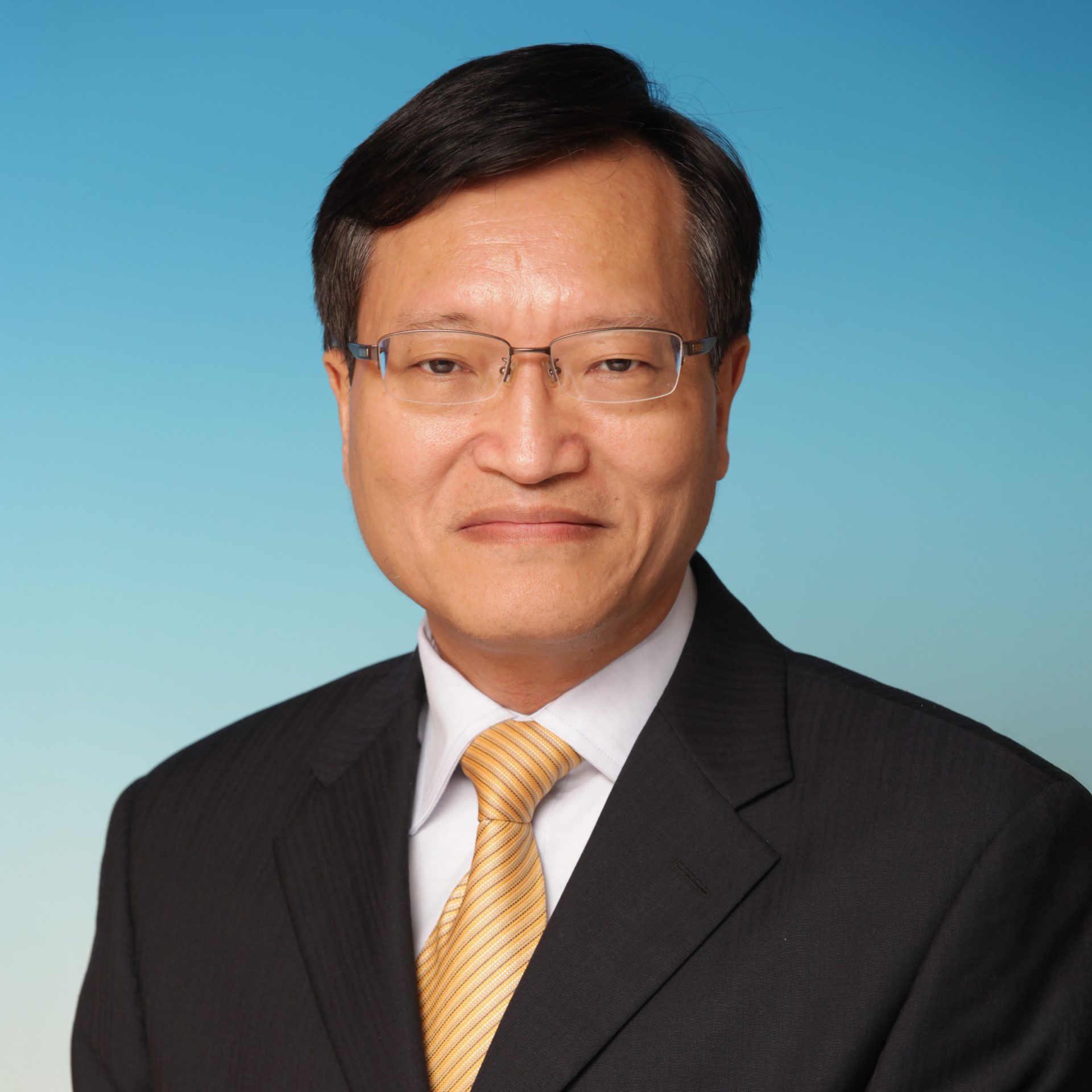 Portrait of Peter Chak wearing glasses, dark suit, and yellow striped tie against blue background.