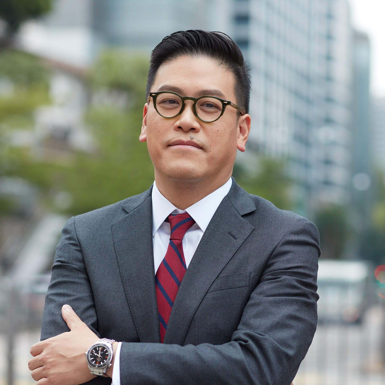 Man in suit and glasses standing confidently with arms crossed outdoors.