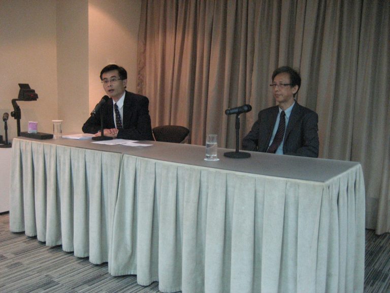 Two men in suits seated at a table with microphones and water glasses.