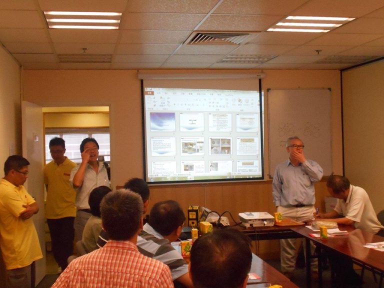 Group of people in meeting room, presenter showing slides on projector screen.