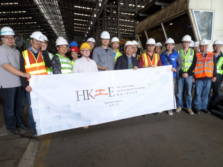 Group of engineers in hard hats holding HKIE Materials Division banner in industrial setting.
