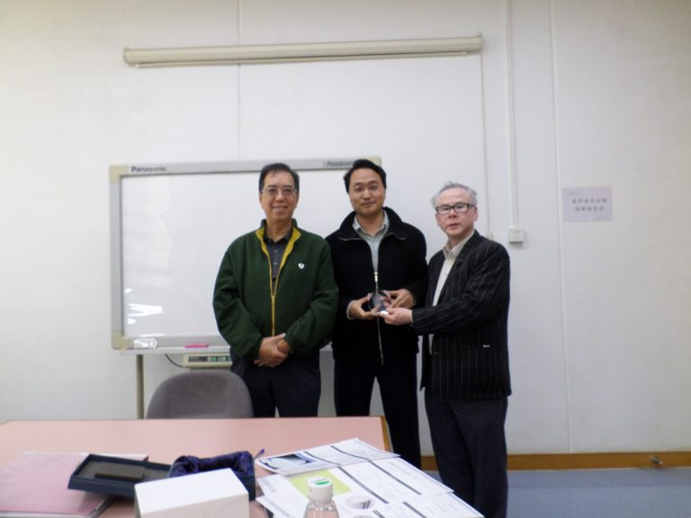 Three men in a classroom setting, one presenting an award to another.
