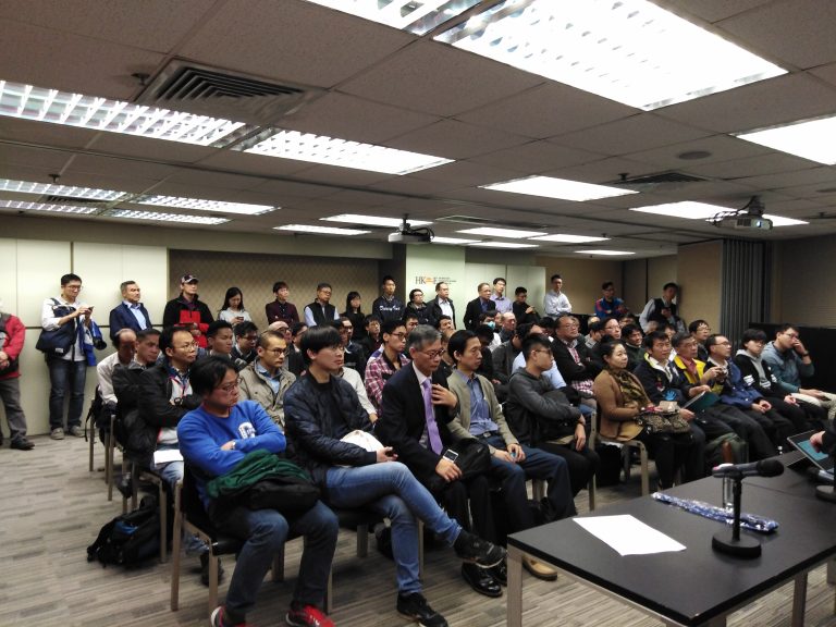 Audience attending a presentation in a conference room with rows of chairs and a speaker's table.