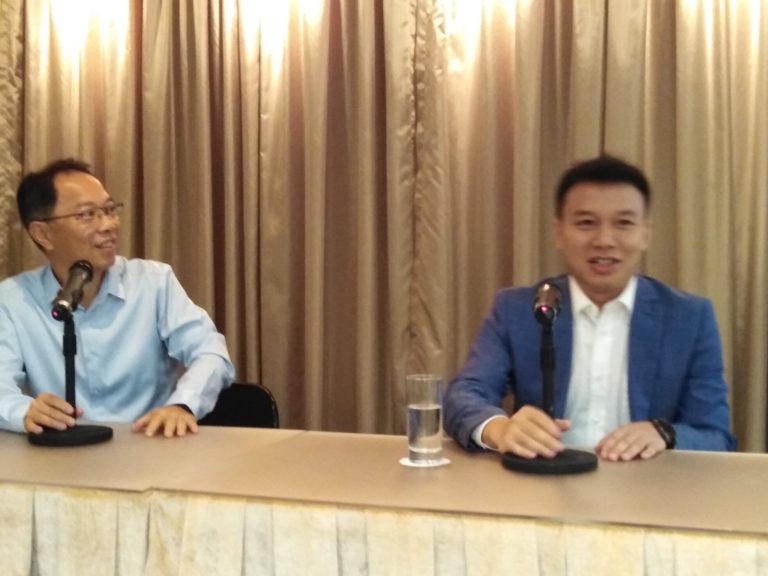 Two men seated at a table with microphones and a glass of water.