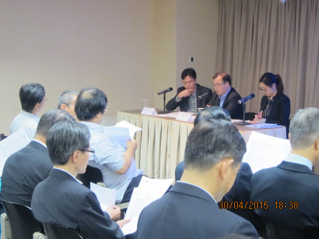 Meeting attendees seated at tables with microphones and documents, viewed from behind.