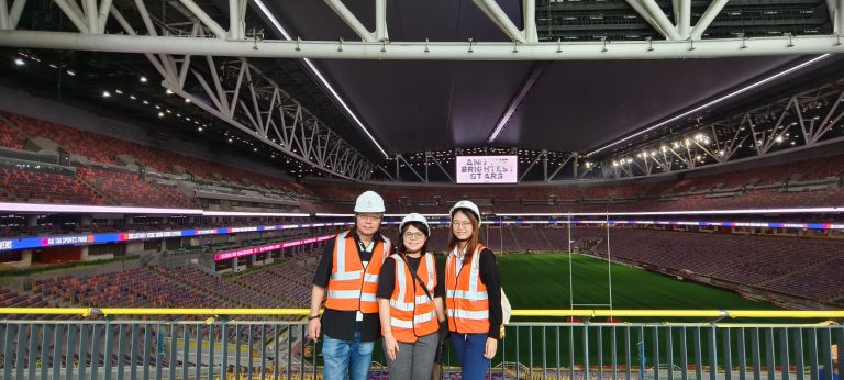 Three people in safety vests and hard hats pose inside a large stadium with rugby field and seating.