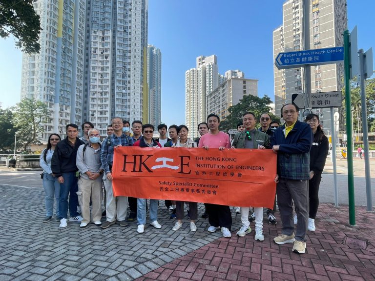 Group of engineers holding HKIE Safety Specialist Committee banner outdoors near high-rise buildings.