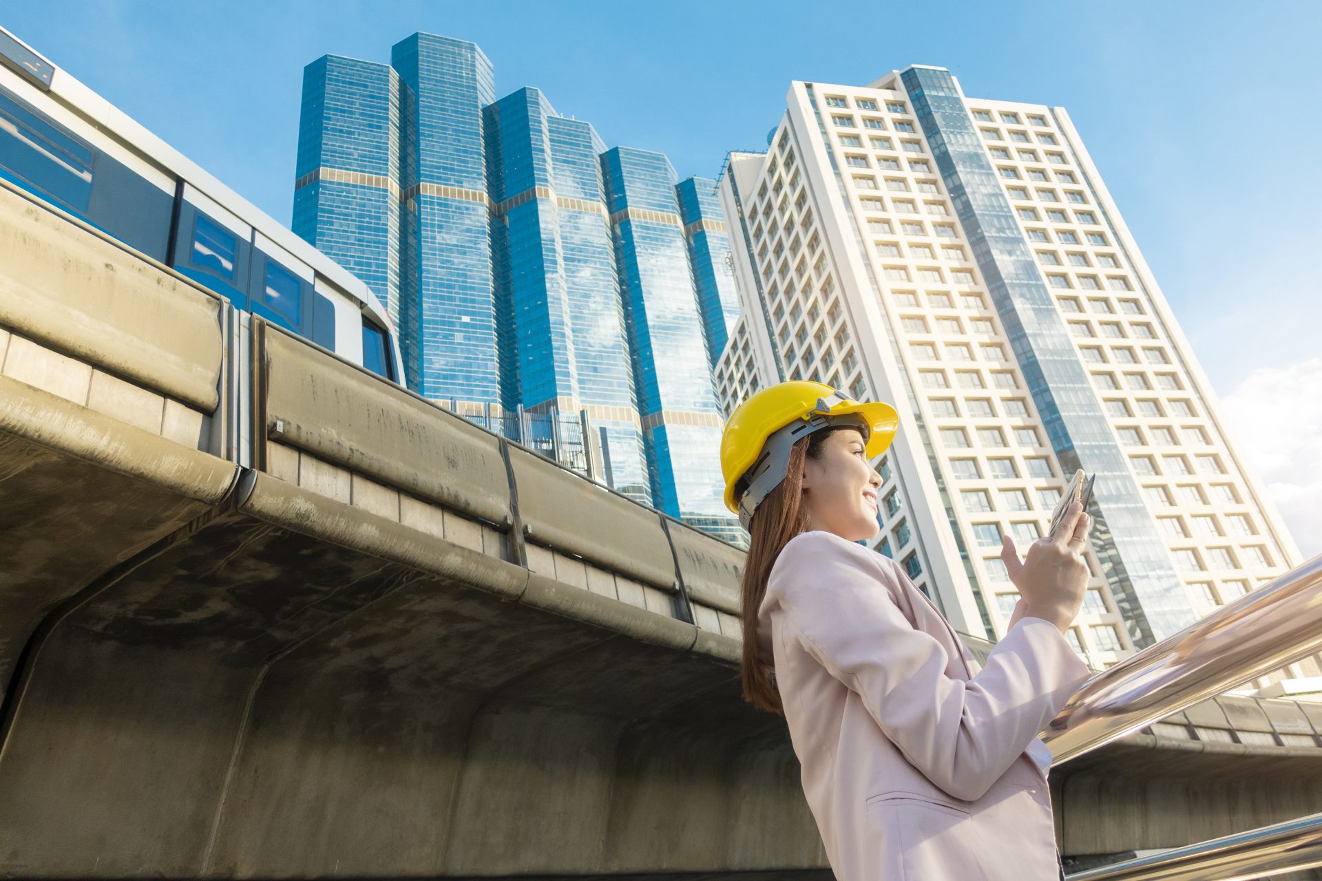 Woman in yellow hard hat using smartphone outdoors near city buildings and elevated train.