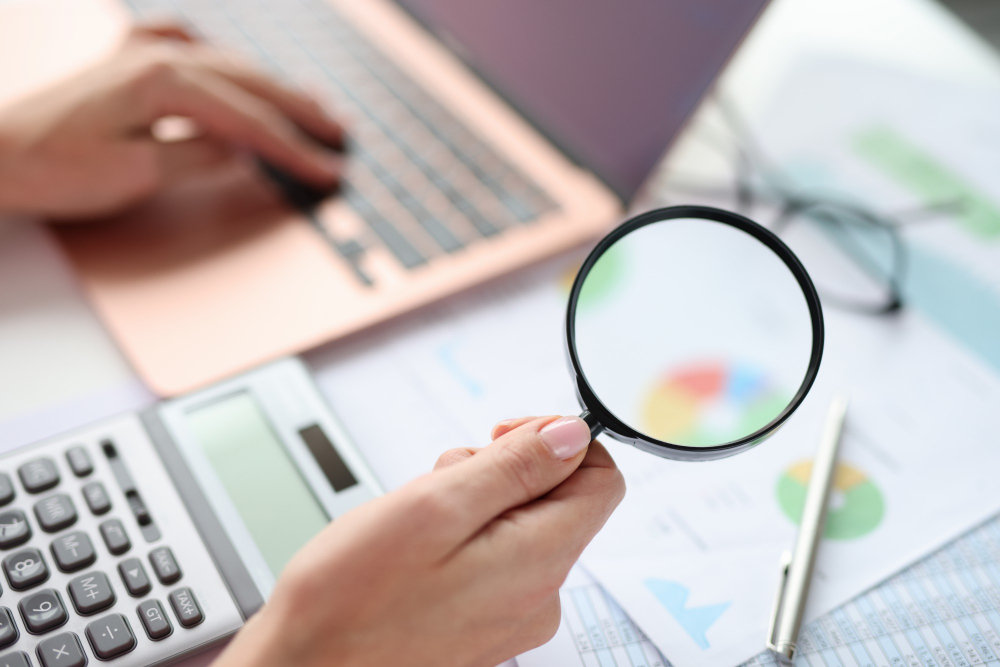 Hand holding magnifying glass over financial documents and laptop