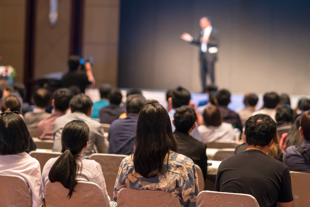 Audience members seated in a conference hall watching a speaker on stage.
