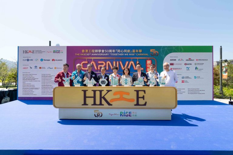 Group of people posing at HKIE 50th Anniversary Carnival event with sponsor logos and engineering-themed props.