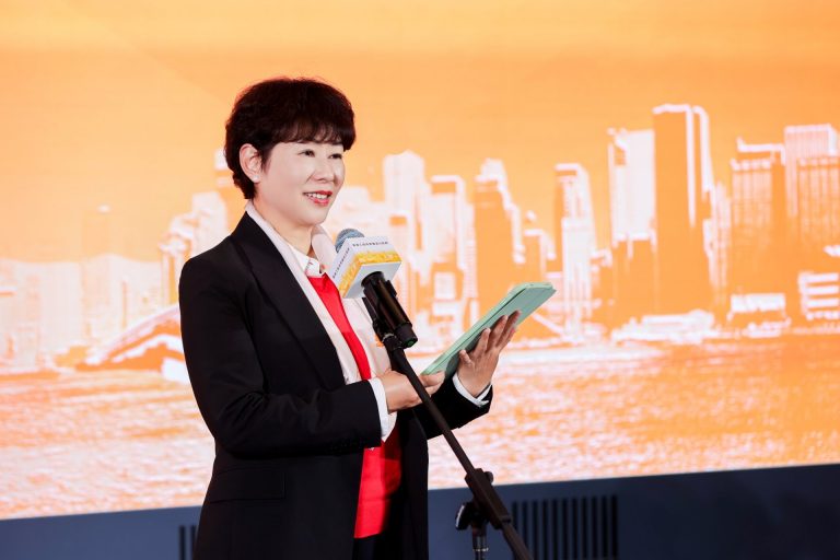 Woman speaking at event with tablet and microphone against orange cityscape backdrop
